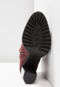 Semelle en caoutchouc noir d'une botte en daim bordeaux avec des crampons à motif, un talon bas et une fermeture éclair latérale, inclinée contre un fond blanc en coin.