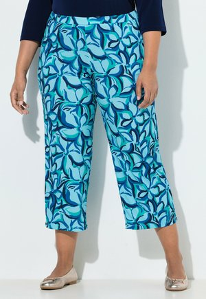 Mujer con pantalones recortados de estampado floral azul intenso y bailarinas metálicas doradas, de pie contra una pared lisa.