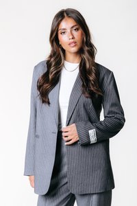 Harmaa pinstripe-takki, jossa on terävät läpät, yksittäinen napitus ja hihassa olevat korostetut tarrat. Yhdistetty valkoiseen yläosaan.