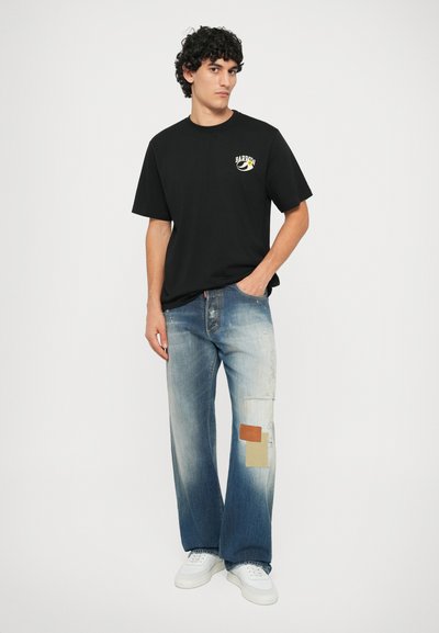 Schwarzes Baumwoll-T-Shirt mit Logo; hellblaue Jeans im weit geschnittenen Stil mit Patchwork- und ausgebleichten Teilen; weiße Sneaker. Einfacher Hintergrund.