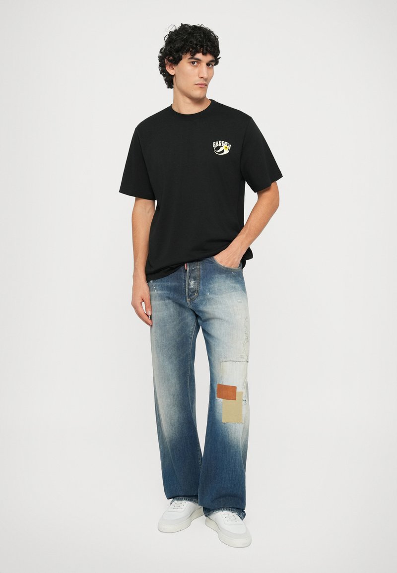 Juoda medvilninė marškinė su logotipu; šviesiai mėlynos džinsinės plačios kelnės su aplikacijomis ir išblukusiomis dalimis; baltos sportbačių. Paprasta fono.