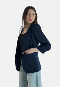 Blazer blu navy con vestibilità su misura, spalle strutturate e maniche arrotolate, abbinato a jeans in denim chiaro. Tessuto morbido e design con apertura frontale.