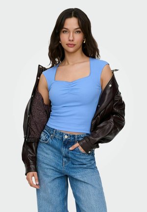 Woman with dark wavy hair wearing a light blue ruched top, high-waisted jeans, and a partially off black leather jacket.