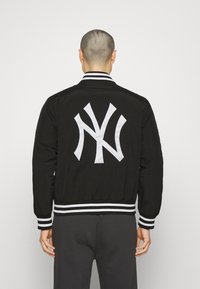 Person wearing black bomber jacket with large white stylized NY logo on back, standing against plain light background.