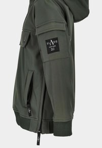 Rękaw ciemnozielonej kurtki z zamkiem błyskawicznym, kieszenią z klapą, ściągaczem oraz czarną prostokątną naszywką z napisem "SLADE COUTURE" na górnej części ramienia.