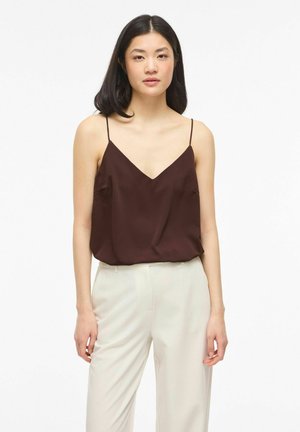 Jeune femme aux cheveux noirs et raides, portant un haut sans manches marron foncé et un pantalon beige clair, debout devant un fond blanc.