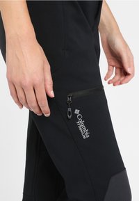 Svarta softshellbyxor med en ficka med dragkedja på vänster lår, med en slät yta och ett Columbia Titanium-logotyp.