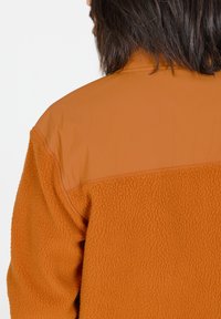Veste en polaire orange avec une base texturée et duveteuse et des épaules lisses de couleur orange plus claire. Présente un détail de couture sur le dos.