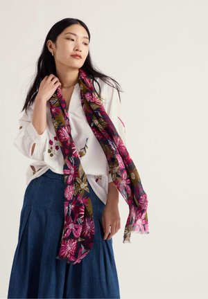 Femme portant un chemisier blanc à fleurs, une jupe plissée bleue et une longue écharpe fleurie rose et verte sur un fond uni.
