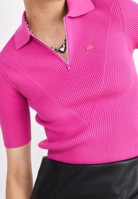 Rosa ribbad polo-tröja med dragkedja i halsen, korta ärmar och subtilt logodetalj på bröstet. Buren med en svart läderkjol.