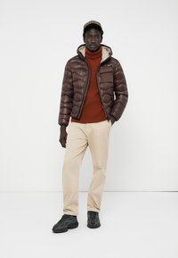 Braune Pufferjacke mit einem cremefarbenen Innenfutter, Brusttasche, hohem Kragen, kombiniert mit einem rostfarbenen Rollkragenpullover, beigen Hosen und schwarzen Sneakern.