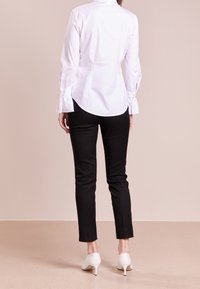Witte blouse met lange mouwen en manchetdetails, gecombineerd met slim-fit zwarte broek en witte hoge hakken, gezien vanuit de achterkant.