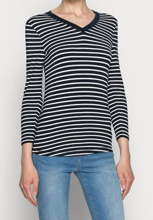 Person iført en sort og hvid langærmet V-hals trøje med vandrette striber og lyseblå jeans, stående mod en ensfarvet baggrund.