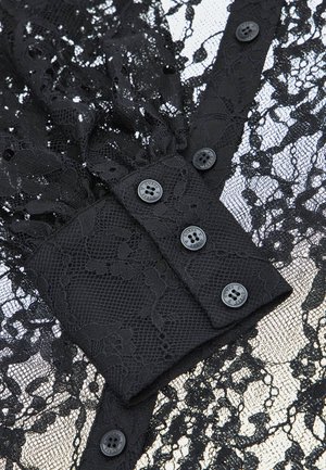Blouse en dentelle noire avec un design texturé, fermeture à boutons et motifs floraux. Le tissu comprend des éléments transparents et opaques.