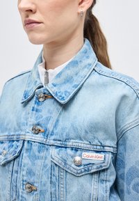Γυναίκα που φοράει ανοιχτό μπλε τζιν μπουφάν με επεξεργασία acid-wash, ασημένια κουμπιά και ετικέτα Calvin Klein στην τσέπη του στήθους, στραμμένη προς τα αριστερά.