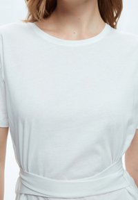 White cotton top featuring a round neckline and short sleeves, with a cinched waist detail and soft, smooth texture.