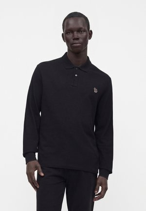 Bărbat purtând un tricou polo negru cu mânecă lungă, cu un mic logo brodat cu zebra multicolor pe piept, stând în fața unui fundal simplu.