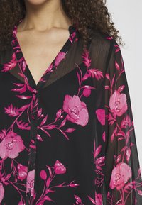 Maglietta nera con motivo floreale rosa, tessuto leggero, scollatura a V e chiusura con bottoni. Presenta maniche trasparenti e dettagli floreali.