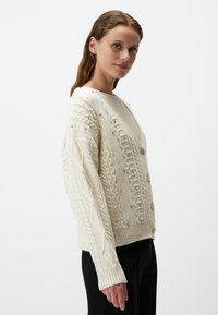 Ivoor gebreide cardigan met kabelpatronen, V-hals en decoratieve zilveren embellissementen langs de voorkant en mouwen. Ribbelzoom.
