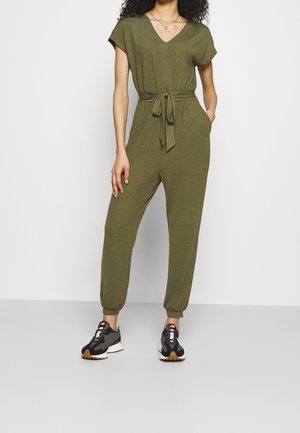 Oliwkowy kombinezon z krótkimi rękawami, dekoltem w kształcie litery V, przewiązanym w talii i zwężającymi się nogawkami. Nosi się go z czarno-białymi butami sportowymi.