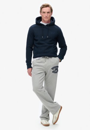 Navy hoodie with drawstrings, front pocket, and gray sweatpants featuring a blue logo. Model wears beige sneakers, standing with one foot forward.