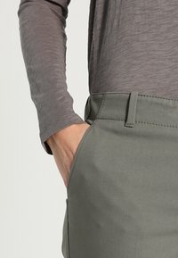 Graues Langarmoberteil kombiniert mit grünen Stoffhosen; die Hosen haben eine glatte Textur und Taschen, sind maßgeschneidert geschnitten und zeigen sichtbare Nahtdetails.