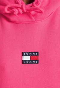 Růžová mikina s měkkou texturou, s vyšitým logem TOMMY JEANS ve modré, bílé a červené barvě. Na kapuci jsou viditelné šňůrky na stažení.