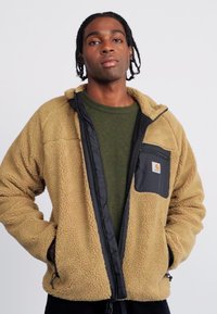 Tan fleecejakke med en sort lynlås og brystlomme; har en blød tekstur og afslappet pasform; båret over en grøn sweater.