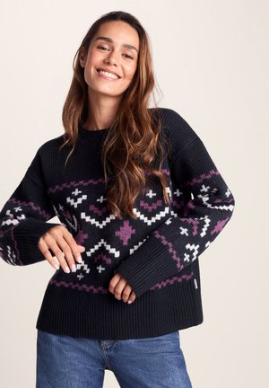 Young woman smiling, wearing a black sweater with white and purple geometric patterns, paired with blue jeans against a plain background.