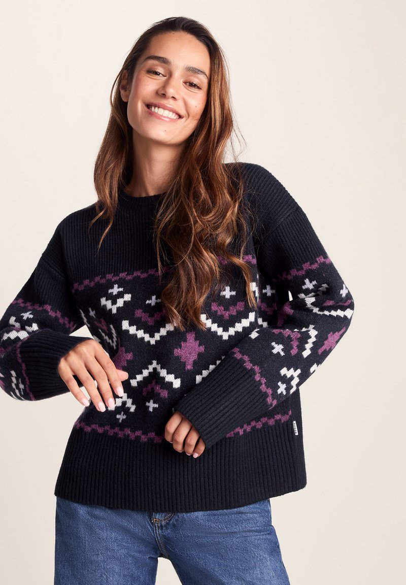 Young woman smiling, wearing a black sweater with white and purple geometric patterns, paired with blue jeans against a plain background.
