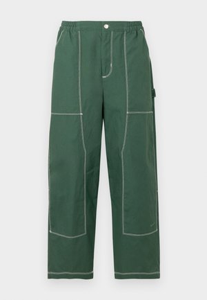 Zielone spodnie o szerokich nogawkach z elastycznym pasem, przednim guzikiem, dużymi widocznymi białymi przeszyciami oraz boczną kieszenią narzędziową na białym tle.