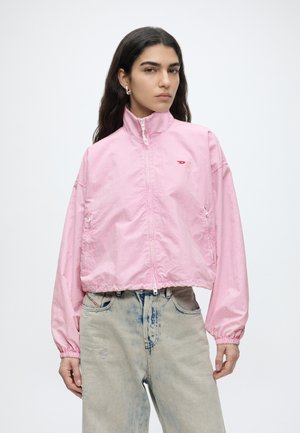 Frau mit langen dunklen Haaren, die eine hellrosa Jacke mit Reißverschluss und ausgebleichte hoch taillierte Jeans trägt, steht vor einem schlichten weißen Hintergrund.