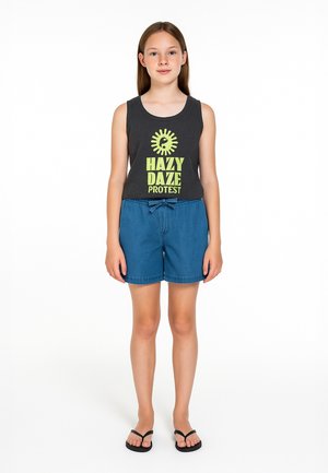 Mladi djevojka stoji, nosi crni top bez rukava sa žutim tekstom "Hazy Daze Protest" i plave traper kratke hlače s vezanjem u struku.