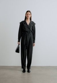Črni kombinezon z oblikovanim ovratnikom, dolgimi rokavi, poudarjenim pasom in širokimi hlačnicami. Dopoplja ga majhna črna torbica in zašiljeni čevlji.