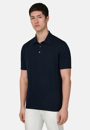 Nuori mies, joka pitää päällään tummansinistä lyhythihaista poolopaitaa ja valkoisia housuja, seisoo yksinkertaisen vaaleanharmaan taustan edessä.