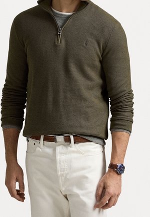 Mężczyzna w oliwkowozielonym swetrze z suwakiem do połowy wysokości, noszący pod nim szarą koszulę, białe spodnie z brązowym paskiem oraz zegarek z niebieską tarczą i brązowym skórzanym paskiem.