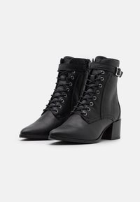 Botas de tobillo de cuero negro con punta afilada, cordones en la parte delantera, cierre de cremallera y un tacón bloque cónico. Cuenta con una correa ajustable con hebilla.