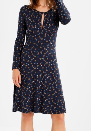 Vrouw draagt een marineblauwe knielange jurk met lange mouwen en kleine meerkleurige bloemenprint, met een keyhole halslijn aan de voorkant.