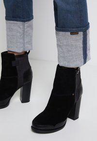 Botines de ante negros con tacón ancho alto, que cuentan con un cierre lateral y un panel texturizado, combinados con unos vaqueros de mezclilla remangados en los puños.