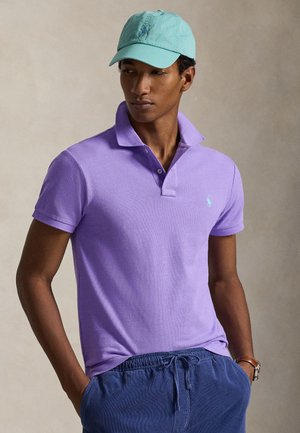 Jeune homme portant un polo lavande avec col relevé, casquette de baseball bleu sarcelle, pantalon bleu à cordon et montre-bracelet marron, mains dans les poches.