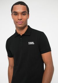 Moški nosi kratkosleeve polo majico črne barve znamke Karl Lagerfeld, stoji ob enobarvni svetlo sivi ozadju.