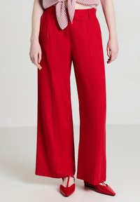 Rote weit geschnittene Hosen aus einem glatten Stoff mit Falten. Getragen mit einem gestreiften Oberteil und passenden roten spitzen Ballerinas, die die kräftige Farbe betonen.