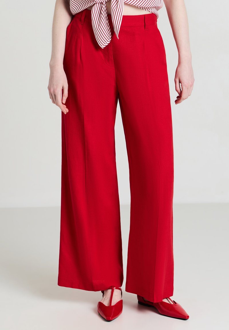 Rote weit geschnittene Hosen aus einem glatten Stoff mit Falten. Getragen mit einem gestreiften Oberteil und passenden roten spitzen Ballerinas, die die kräftige Farbe betonen.