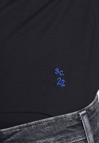 Zwart stof met fijne textuur, met blauwe geborduurde tekst "SC. 22" aan de onderkant. Vergezeld van een grijze spijkerstof tailleband.