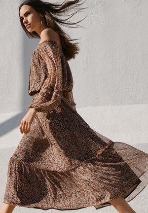 Žena kráčajúca v prúdiacich hnedých kvetovaných šatách bez ramien, vlasy vlajú, na pozadí bielej textúrovanej steny na slnečnom svetle.