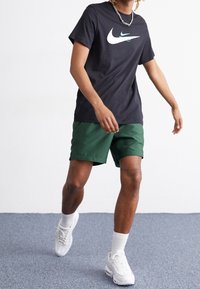 En person går inomhus iförd en svart Nike-t-shirt, gröna shorts, vita strumpor och vita sneakers på en blå matta.