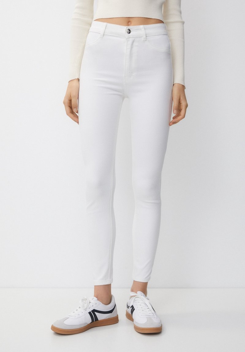 Femme portant un jean skinny blanc taille haute, un top blanc à manches longues court et des baskets blanches avec semelles noires et marron, debout sur un sol blanc.