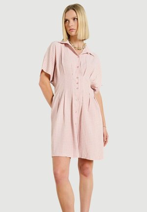 Frau trägt ein hellrosa, kurzärmeliges Kleid mit Knopfleiste und einem dezenten Karomuster sowie eine auffällige goldene Gliederkette.