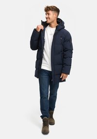Chaqueta acolchada de color azul marino con capucha, con bolsillos con cremallera, cremallera frontal y un ajuste relajado. Combinada con jeans de mezclilla y botas marrones.
