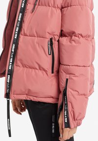 Roze pufferjack met een gewatteerd ontwerp, met zwarte ritsen en contrasterende tape langs de ritsen. De stof lijkt glad en lichtgewicht.